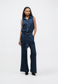 Denim jumpsuit with sleeveless design, collared neck, front buttons, elastic waist, and wide legs. Dark blue fabric with a smooth texture.