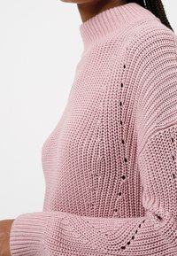 Jasnoróżowy sweter dziewiarski z wysokim kołnierzem, teksturowanym materiałem i otwartymi detalami ściegów na rękawach. Miękki, oddychający materiał.