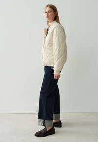 Quilted creme Bomberjacke mit gerippten Bündchen, kombiniert mit dunkelblauen weit geschnittenen Jeans mit umgeschlagenen Säumen und braunen Slippern.