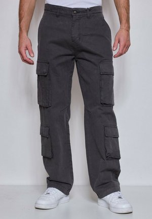 Homme portant un pantalon cargo gris foncé avec plusieurs poches à rabat et des baskets blanches, debout devant un fond clair uni.