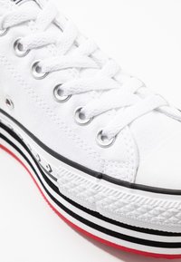 Vita canvas sneakers med en texturerad gummitå, silverfärgade öglor och en distinkt svart och röd randig sula. Har vita snörningar.