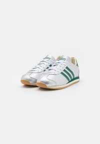 White and silver Adidas sneakers with green stripes and gum soles, viewed from the front on a white background.