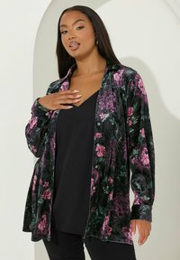 Giacca camicia in velluto nero con motivo floreale, caratterizzata da fiori rosa e verdi, design a un bottone, maniche lunghe e vestibilità rilassata sopra una maglietta nera.
