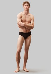 Bañador negro de tejido suave, con una cinturilla ancha. El modelo posa con los brazos cruzados, mostrando un físico en forma y pies descalzos.