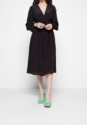 Robe noire à manches longues avec un décolleté en V et une taille froncée, présentant un design fluide. Associée à des talons ouverts verts et des bagues bleues et orange.