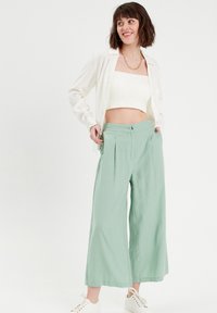 Pantalons larges en lin vert clair avec des plis à l'avant, associés à un top court blanc et des baskets blanches, sur un fond uni.