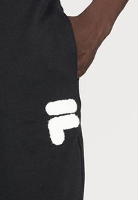 Pantaloni della tuta neri con un logo bianco testurizzato sul lato sinistro. Il tessuto sembra morbido con una leggera lucentezza, evidenziando un design casual.