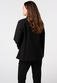 Zwarte op maat gemaakte blazer met een gestructureerde pasvorm, gladde stof en lange mouwen. Voorzien van een achternaad en minimale hardware-accenten.