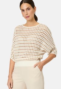 Cremfarbener, offen gestrickter Pullover mit lockerer Passform, Rundhalsausschnitt und geripptem Saum. Die Textur ist durchsichtig und zeigt die darunterliegende Haut.