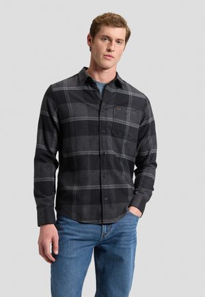Giovane uomo che indossa una camicia a quadri nera e grigia con bottoni e jeans blu, in piedi con una mano in tasca davanti a uno sfondo semplice.