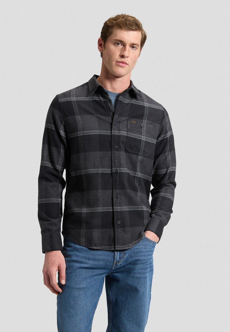 Jeune homme portant une chemise à carreaux noire et grise boutonnée et un jean bleu, debout avec une main dans la poche devant un fond uni.