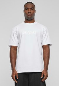 White cotton t-shirt with short sleeves, featuring the word "KAN" in a reflective print on the front, paired with black trousers.