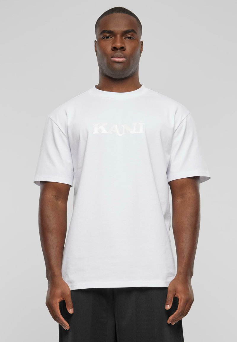 White cotton t-shirt with short sleeves, featuring the word "KAN" in a reflective print on the front, paired with black trousers.