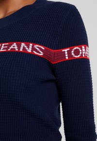 Maglione in maglia waffle blu navy con striscia orizzontale rossa e bianca sul petto e sul braccio superiore, con testo parzialmente visibile.