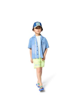 Jeune garçon portant une casquette bleue, une chemise en denim à manches courtes, un short vert clair et des sandales bleues, marchant sur un fond blanc.