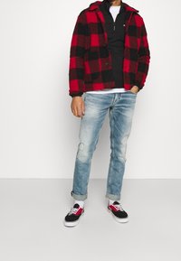 Veste en polaire à carreaux rouges et noirs, sweat-shirt zippé noir, jean bleu clair déchiré et baskets noires avec des accents rouges.