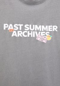 Grå skjorte med hvid tekst "PAST SUMMER ARCHIVES" og tre små klistermærker: orange prisskilt, pink Lissabon transportbillet og orange skive klistermærke.