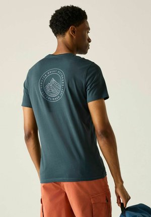 Joven con camiseta color azul verdoso con logo de montaña en la espalda, pantalones cortos cargo naranjas, sujetando la asa de una mochila azul a un lado.