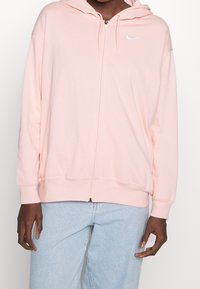 Osoba v růžové mikině na zip značky Nike a světle modrých džínách stojící uvolněně s pažemi podél těla na bílém pozadí.