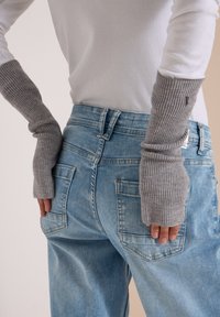 Personne portant un haut blanc, des gants gris tricotés sans doigts et un jean bleu clair ample, les mains reposant près des poches arrière.
