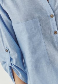 Camicia button-up azzurro chiaro realizzata in tessuto morbido, con taschino sul petto e maniche arrotolate con dettagli a bottoni.