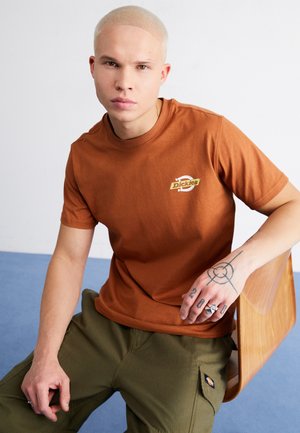 Camiseta de algodón marrón con un pequeño logotipo beige en el pecho, combinada con pantalones cargo verde oliva. Textura suave, corte informal. Silla de madera al fondo.