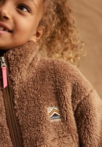 Giacca in pile marrone con colletto alto, dotata di zip e un tirante rosa. Una patch mostra il marchio "Superdry Mountain Sport."