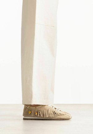 Beige mocassin-stijl sneakers met franjes, witte veters en kleine decoratieve kraaltjes, gedragen met lichtgekleurde wijde broekspijpen.