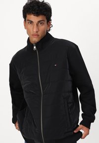 Black zip-up jacket with a padded front and solid black sleeves. Features a high collar, ribbed cuffs, and a small logo on the chest.