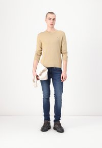 Beige strikkegenser, mørkeblå jeans og svarte joggesko. Modell som holder et hvitt stoff. Enkelt bakgrunn, medium langt hår, nøytral uttrykk.