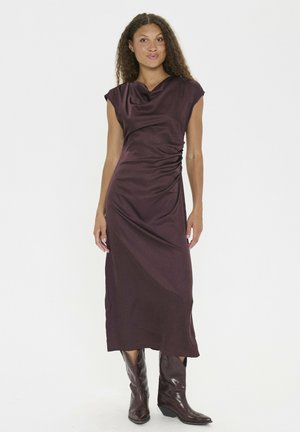 Bordeauxfarbenes, ärmelloses Kleid mit drapiertem Ausschnitt und gerafftem Seitendetail; glatte, satinähnliche Textur; getragen mit braunen Leder-Cowboy-Stiefeln.