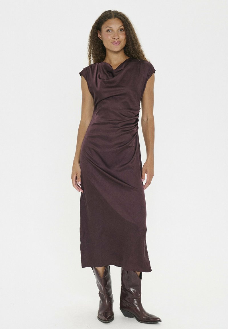Bordeauxfarbenes, ärmelloses Kleid mit drapiertem Ausschnitt und gerafftem Seitendetail; glatte, satinähnliche Textur; getragen mit braunen Leder-Cowboy-Stiefeln.