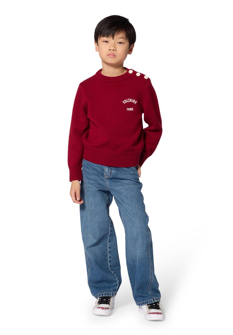 Αγόρι που στέκεται φορώντας κόκκινο πουλόβερ με λευκά κουμπιά στον ώμο, μπλε τζιν και λευκά αθλητικά παπούτσια σε λευκό φόντο.