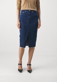 Mujer vistiendo una falda de mezclilla azul oscura de longitud media con una abertura frontal, una blusa de manga larga color crema y tacones negros puntiagudos con correas en los tobillos.