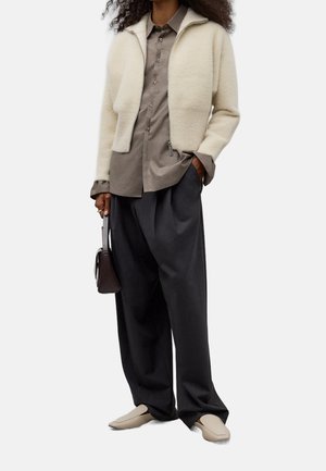 Veste zippée crème duveteuse portée sur une chemise grise boutonnée, associée à un pantalon gris ample et des chaussures beige à enfiler, tenant un petit sac à main sombre.