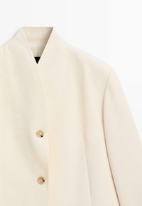 Off-white blazer med høj hals og to beige knapper foran, vist mod en ensfarvet hvid baggrund.