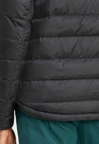 Chaqueta de plumas negra con diseño acolchado y una tela exterior suave y brillante. El dobladillo de la chaqueta es ligeramente curvado. El fondo incluye tela color verde azulado.