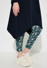 Tunic blu navy con tessuto testurizzato e orlo asimmetrico, abbinata a leggings floreali turchesi e sneakers bianche. Inquadratura ravvicinata della parte inferiore del corpo.