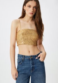 Złota, cekinowa bluzka typu crop top o dopasowanym kroju, na ramiączkach spaghetti i fakturowanej powierzchni, zestawiona z jeansami z wysokim stanem w niebieskim kolorze.