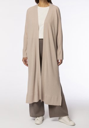 Kobieta w długim beżowym prążkowanym kardiganie na białej bluzce i szerokich brązowych spodniach oraz białych trampkach, stojąca na białym tle.