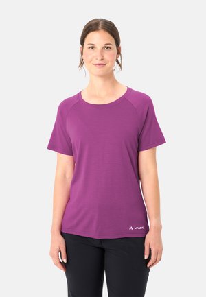 Frau trägt ein lila kurzärmliges T-Shirt und schwarze Hosen, steht und blickt nach vorne vor einem weißen Hintergrund.