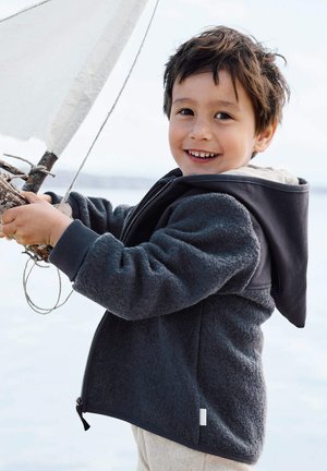 Lächelndes Kind in dunkler Fleecejacke, das ein kleines Segelboot nahe dem ruhigen Wasser unter einem strahlend blauen Himmel hält.