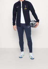 Giacca con zip della marina con logo e branding, abbinata a pantaloni coordinati e scarpe bianche con dettagli blu e verdi. Tiene un pallone da calcio.