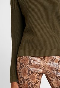 Oliivinvihreä ribbineulottu pusero, jossa on teksturoitu pinta, yhdistettynä istuviin ruskean ja beigen sävyisiin käärmekuvioisiin leggingsseihin, joissa on sileä viimeistely.