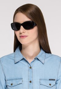 Lunettes de soleil noires avec une monture large et courbée ; finition brillante ; portées avec une chemise en denim bleu clair comportant des poches poitrine et des détails de boutons.