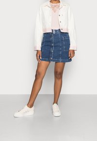 Weißejacke aus Denim mit cropped Schnitt und gestreiften Bündchen, hellrosa Top, blaue A-Linie Jeansrock mit knöpfbarem Frontverschluss und hellen Sneakern mit Akzenten.