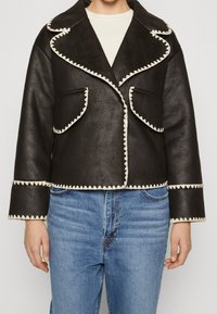 Zwart leren jack met witte golvende afwerking, voorzien van een brede kraag, voorzakken en een cropped ontwerp, gecombineerd met blauwe jeans.