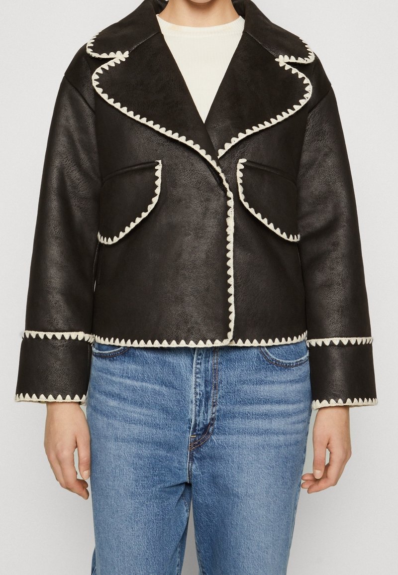 Zwart leren jack met witte golvende afwerking, voorzien van een brede kraag, voorzakken en een cropped ontwerp, gecombineerd met blauwe jeans.