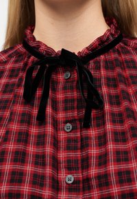 Γυναίκα που φοράει κόκκινο και μαύρο πουκάμισο με καρό σχέδιο, με μαύρη βελούδινη κορδέλα δεμένη σε φιόγκο στη στρογγυλεμένη λαιμόκοψη με βολάν και μαύρα κουμπιά στο μπροστινό μέρος.