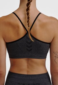 Kobieta z zaplecionymi włosami, ubrana w ciemnoszary sportowy biustonosz z cienkimi ramiączkami i dopasowane legginsy z wysokim stanem, pokazująca tatuaż jaszczurki na ramieniu.